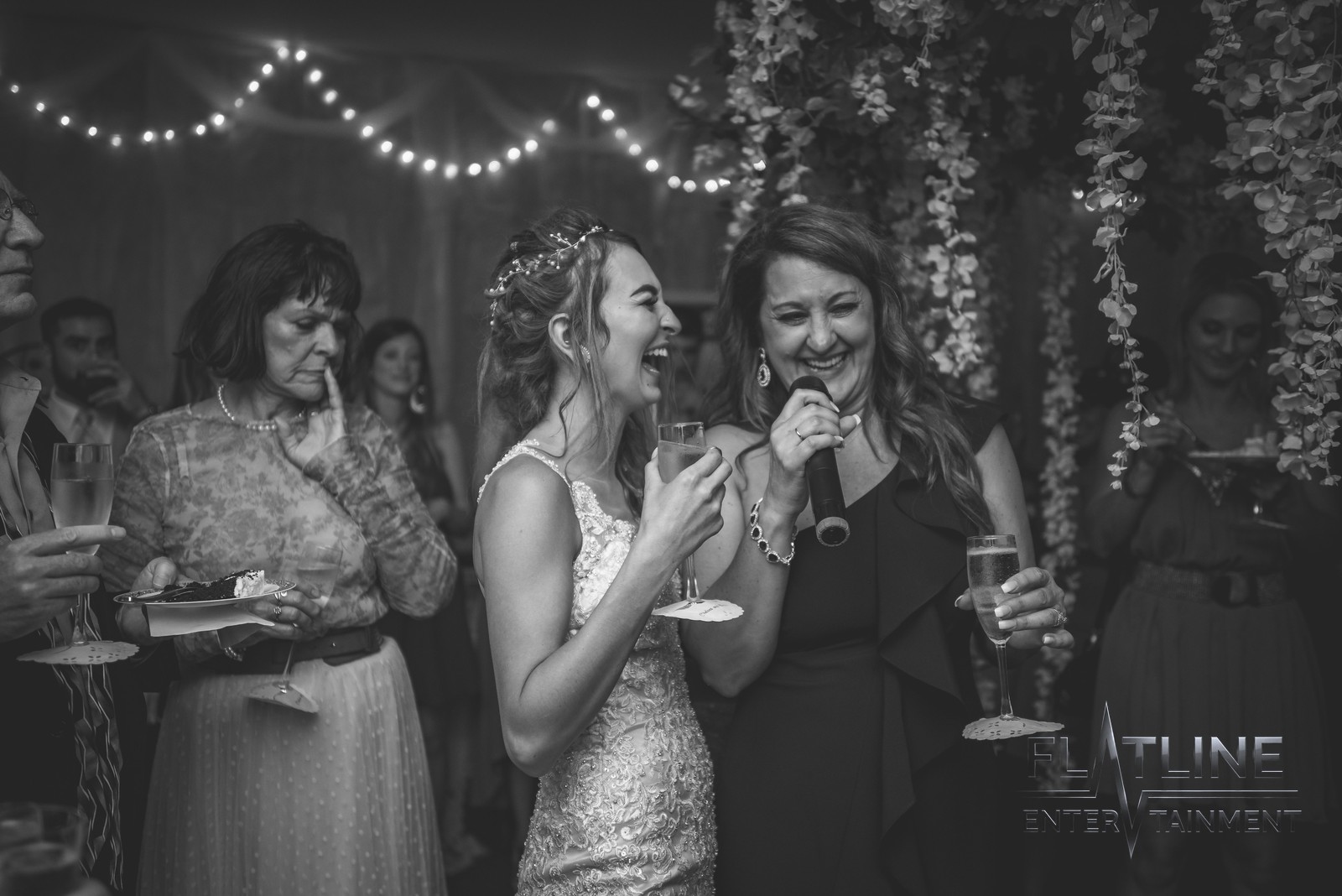 Black-and-white photo of a laughing bride beside a guest speaking into a microphone at a wedding reception