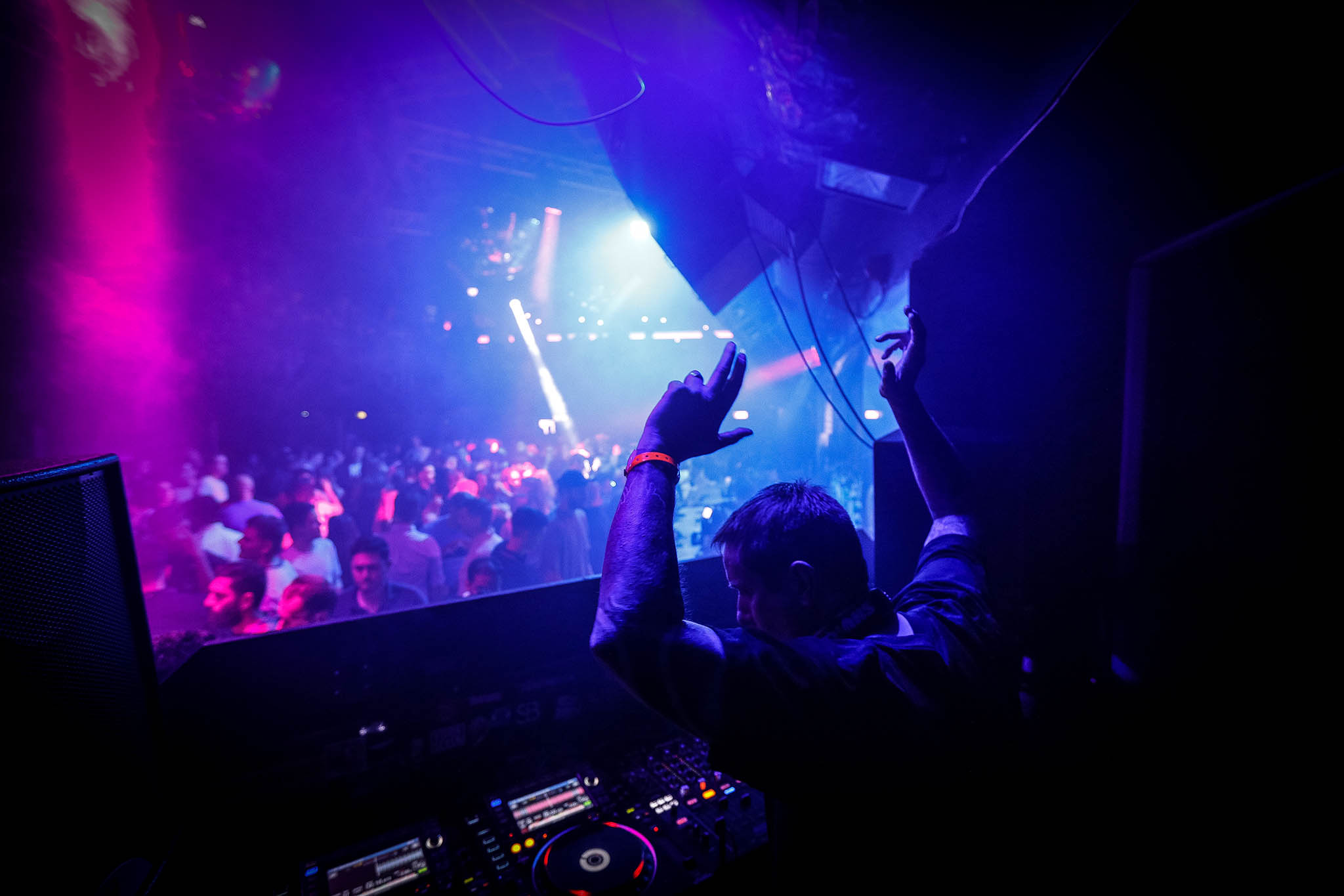 A DJ with hands raised over a packed, light-filled dance floor