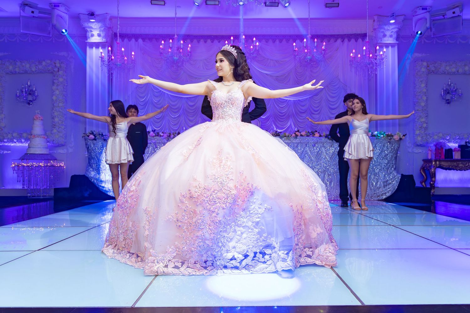 A quinceanera in a blush ballgown dancing with her court in a decorated ballroom